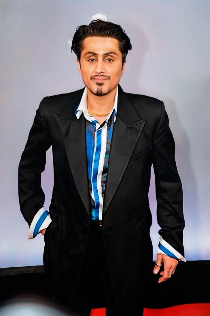 Samuel Sohebi bei den Reality Awards 2025 in Bonn, trägt einen schwarzen Anzug mit auffälligem blau-gestreiften Hemd, frontal zur Kamera stehend in einer selbstbewussten Pose vor neutralem Hintergrund.