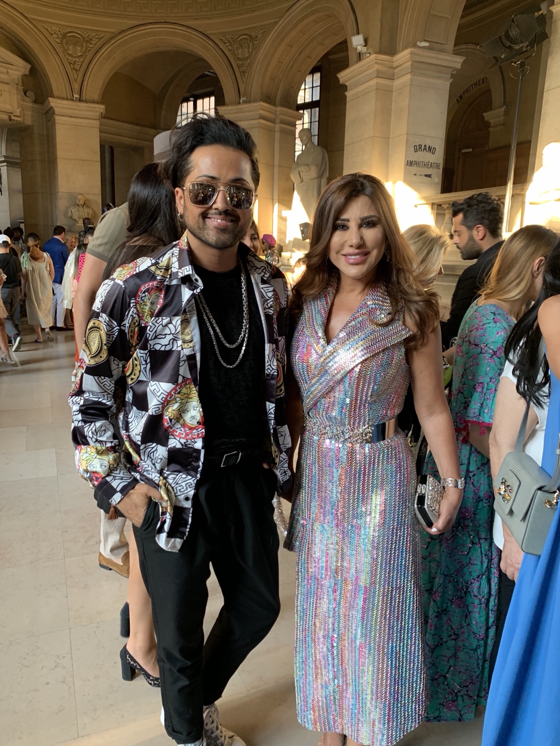 Najwa Karam in einem bunt schimmernden Metallic-Wickelkleid von Rami Kadi neben Samuel Sohebi mit gemustertem Hemd und Sonnenbrille in einer hohen Halle mit Rundbögen.